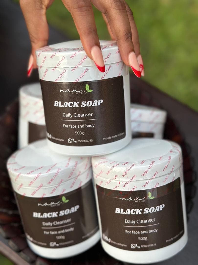 A hand resting on a stack of NAX African Black Soap jars, outdoors